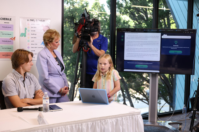 A young girl speaks at a table with a laptop while a woman, a seated boy, and a cameraman watch, with a large screen displaying text behind them.